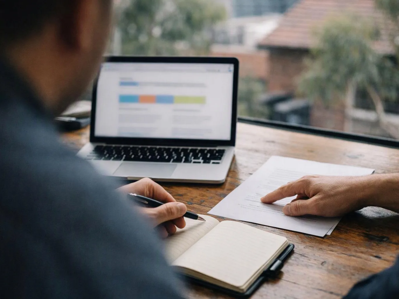 Two people review a document at a wooden table beside a laptop and notebook