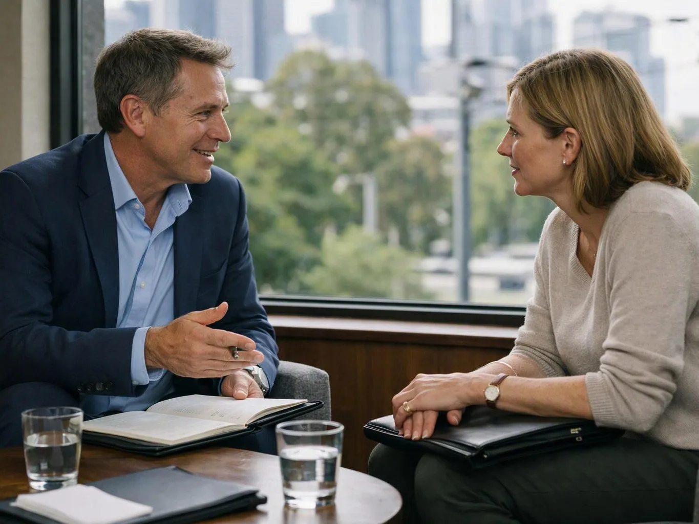 Two people sit by a window, talking with notebooks and water glasses on a table